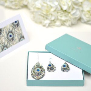 May include: A teal gift box with a white lid containing a peacock feather necklace and earrings. The necklace and earrings are made of resin and feature a peacock feather design with blue, green, and gold colors. The box is sitting on a white surface.