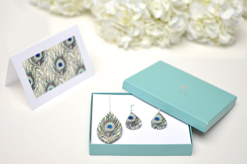 May include: A teal gift box with a white lid containing a peacock feather necklace and earrings. The necklace and earrings are made of resin and feature a peacock feather design with blue, green, and gold colors. The box is sitting on a white surface.