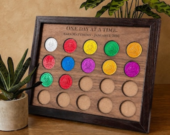 May include: A dark wood display board featuring the engraved text "ONE DAY AT A TIME." and "SARA MATTHEWS - JANUARY 1, 2010." The board contains multiple circular cutouts, some of which hold colorful sobriety coins in silver, red, yellow, green, orange, blue, and purple.