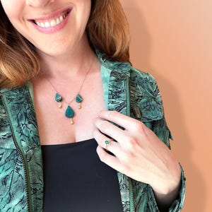 May include: A woman wearing a green patterned jacket with a gold necklace featuring three teardrop-shaped green stones. She is also wearing a ring with a green stone on her right hand.