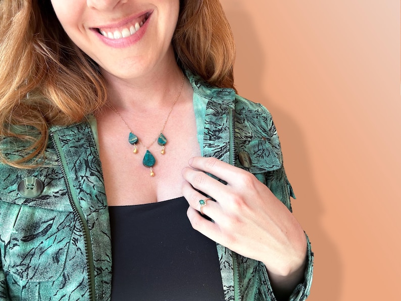 May include: A woman wearing a green patterned jacket with a gold necklace featuring three teardrop-shaped green stones. She is also wearing a ring with a green stone on her right hand.