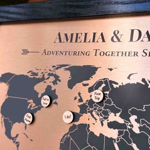 Peut inclure: Une carte du monde de couleur cuivre avec des contours noirs des continents. La carte est encadr&eacute;e de bois noir et comporte quatre marqueurs ronds en bois avec du texte qui dit "First Home", "Fell in love", "I do!" et "First Trip".