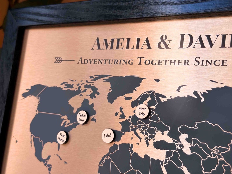 Peut inclure: Une carte du monde de couleur cuivre avec des contours noirs des continents. La carte est encadr&eacute;e de bois noir et comporte quatre marqueurs ronds en bois avec du texte qui dit "First Home", "Fell in love", "I do!" et "First Trip".