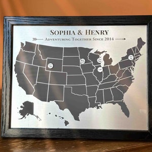 K&ouml;nnte beinhalten: Eine schwarze gerahmte Karte der Vereinigten Staaten mit wei&szlig;en Stecknadeln, die Orte markieren. Die Karte tr&auml;gt den Titel "Sophia & Henry Adventuring Together Since 2014".