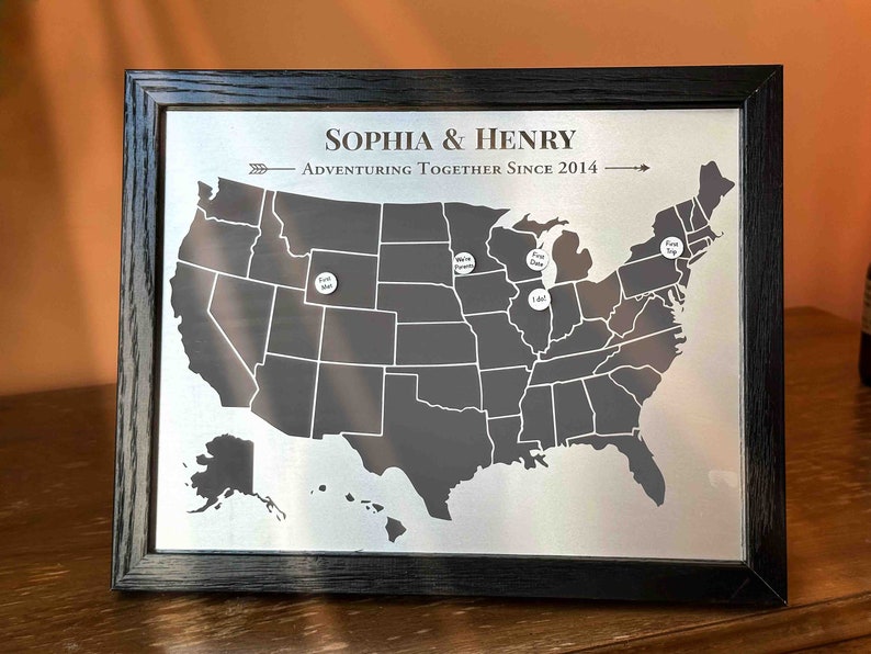 K&ouml;nnte beinhalten: Eine schwarze gerahmte Karte der Vereinigten Staaten mit wei&szlig;en Stecknadeln, die Orte markieren. Die Karte tr&auml;gt den Titel "Sophia & Henry Adventuring Together Since 2014".