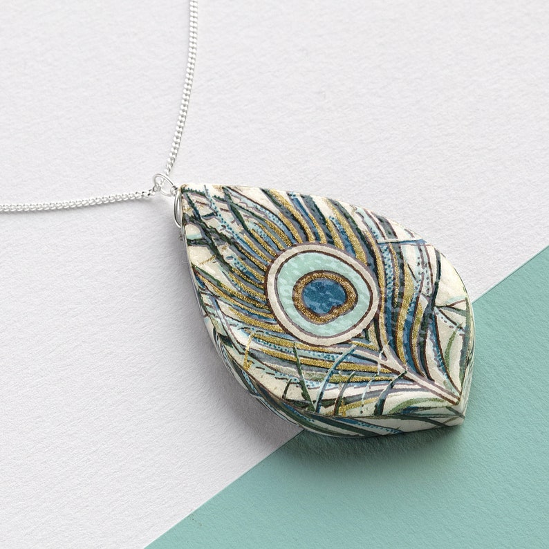 May include: A silver chain necklace with a teardrop-shaped pendant featuring a peacock feather design. The pendant is white with blue, green, and gold accents.