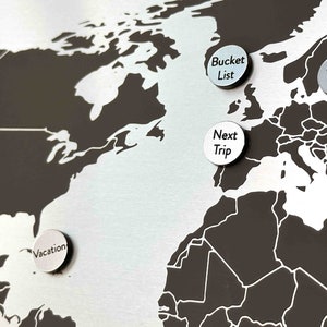 K&ouml;nnte beinhalten: Eine Weltkarte mit silbernen Stecknadeln mit Text darauf. Die Stecknadeln sind auf Nordamerika, Europa und Afrika platziert. Die Stecknadeln zeigen "Vacation", "Bucket List", "Next Trip" und "Fell in love".