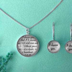 May include: Silver necklace and earrings with a round pendant featuring the text "I choose you. And I'll choose you over and over and over. Without pause, without a doubt, in a heartbeat. 7.21.18" and the earrings each say "I choose you."