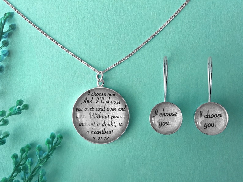 May include: Silver necklace and earrings with a round pendant featuring the text "I choose you. And I'll choose you over and over and over. Without pause, without a doubt, in a heartbeat. 7.21.18" and the earrings each say "I choose you."