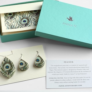 May include: Peacock feather earrings made from Italian paper, in a teal gift box with a white card that says "ANNA V." and "PEACOCK". The card describes the earrings as a symbol of renewal and immortality. The earrings are sterling silver and come with a silica pack. The card also says "PAPER-ANNIVERSARY.COM".