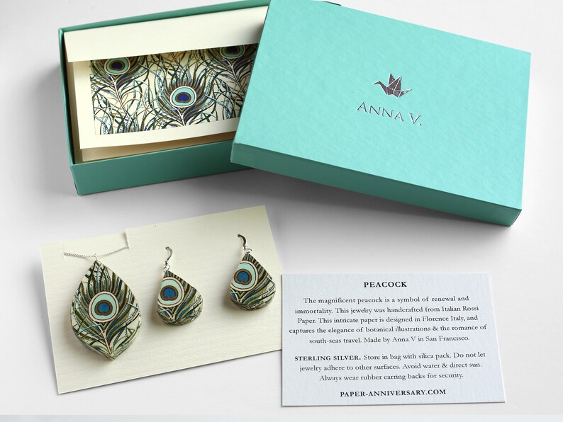 May include: Peacock feather earrings made from Italian paper, in a teal gift box with a white card that says "ANNA V." and "PEACOCK". The card describes the earrings as a symbol of renewal and immortality. The earrings are sterling silver and come with a silica pack. The card also says "PAPER-ANNIVERSARY.COM".