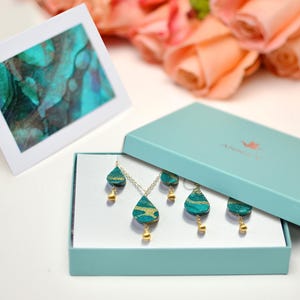May include: A teal and gold necklace and earrings set in a teal gift box. The jewelry features a teardrop design with gold accents. The box has the word "ANNIKA" printed on the top.