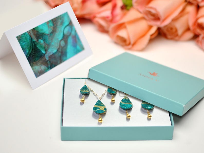 May include: A teal and gold necklace and earrings set in a teal gift box. The jewelry features a teardrop design with gold accents. The box has the word "ANNIKA" printed on the top.