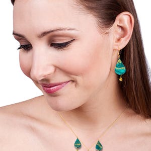 May include: A gold chain necklace with three teardrop-shaped pendants. Each pendant is made of green and gold patterned glass with a small gold bead hanging from the bottom. The necklace is worn by a person with brown hair.