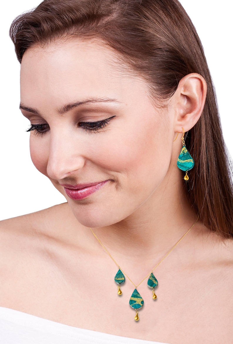 May include: A gold chain necklace with three teardrop-shaped pendants. Each pendant is made of green and gold patterned glass with a small gold bead hanging from the bottom. The necklace is worn by a person with brown hair.