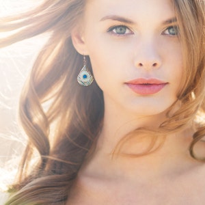 May include: A woman with long, wavy blonde hair wearing a pair of blue and green peacock feather earrings. The earrings are teardrop shaped and have a gold setting.