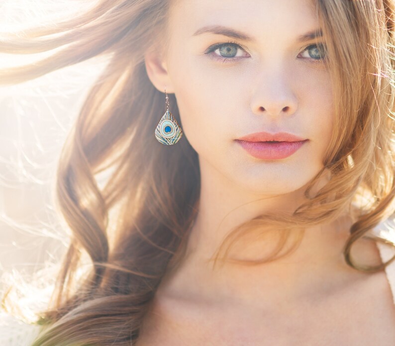 May include: A woman with long, wavy blonde hair wearing a pair of blue and green peacock feather earrings. The earrings are teardrop shaped and have a gold setting.