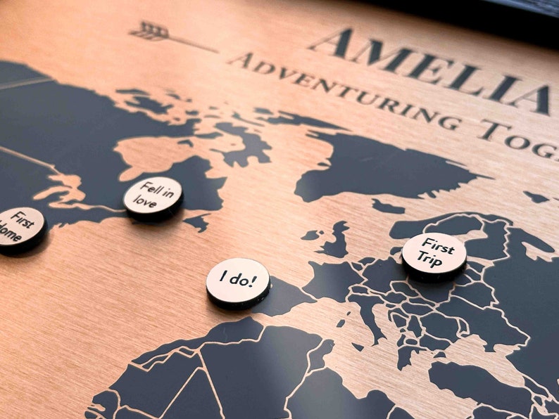 Peut inclure: Une carte du monde en bois avec des continents noirs et un fond couleur cuivre. La carte est intitul&eacute;e "Amelia Adventuring Together". Il y a quatre &eacute;pingles rondes sur la carte avec le texte suivant : "First Home", "Fell in Love", "I do!" et "First Trip".