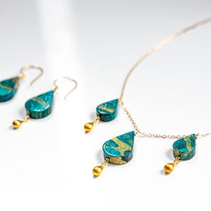 May include: A gold chain necklace with two teardrop-shaped pendants. The pendants are a deep teal blue with gold accents. The necklace also comes with a pair of matching earrings.