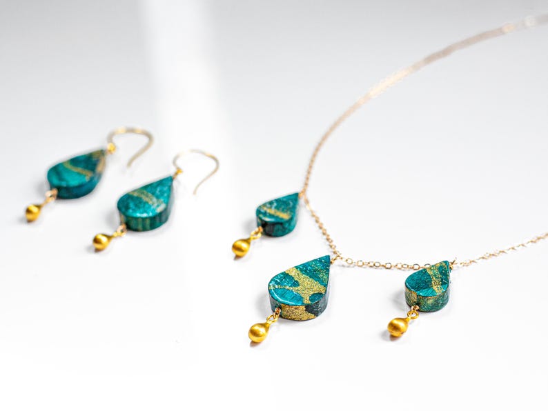 May include: A gold chain necklace with two teardrop-shaped pendants. The pendants are a deep teal blue with gold accents. The necklace also comes with a pair of matching earrings.