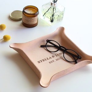 May include: A light pink leather tray with rounded corners, holding a pair of black-framed glasses. The tray is embossed with the text "STELLA & JAMES EST. 2015". A candle and diffuser are in the background.