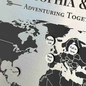 K&ouml;nnte beinhalten: Eine schwarz-wei&szlig;e Weltkarte mit silbernen Stecknadeln. Die Stecknadeln haben Text darauf, der "First Home", "Vacation", "Bucket List", "Fell in Love" und "Next Trip" lautet. Die Karte befindet sich auf einem silbernen Hintergrund mit dem Text "& H ADVENTURING TOGETHER" oben.