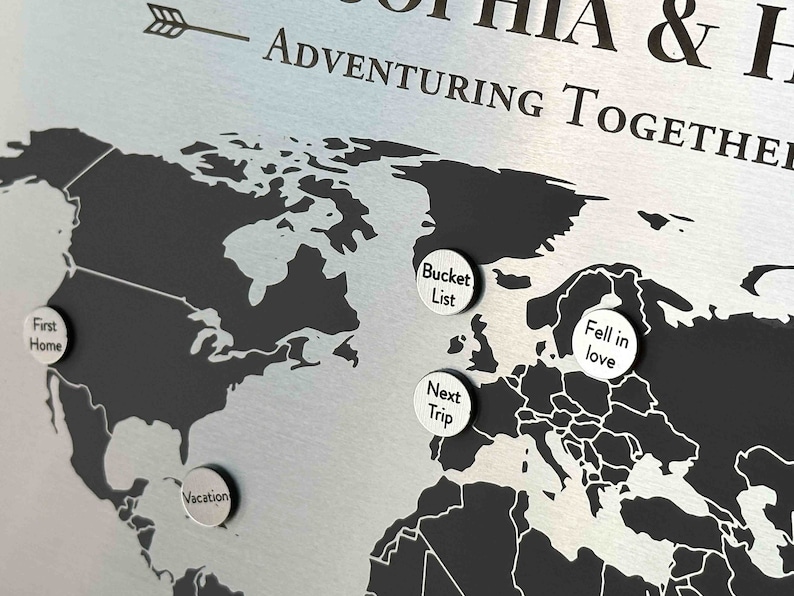 K&ouml;nnte beinhalten: Eine schwarz-wei&szlig;e Weltkarte mit silbernen Stecknadeln. Die Stecknadeln haben Text darauf, der "First Home", "Vacation", "Bucket List", "Fell in Love" und "Next Trip" lautet. Die Karte befindet sich auf einem silbernen Hintergrund mit dem Text "& H ADVENTURING TOGETHER" oben.
