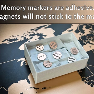 Peut inclure: Une bo&icirc;te blanche contenant une vari&eacute;t&eacute; de marqueurs de m&eacute;moire adh&eacute;sifs ronds. Les marqueurs pr&eacute;sentent diff&eacute;rentes phrases, notamment "Next Trip", "First Home", "We're Parents", "First Date", "Will You?", "Top I" et "boow Lauos". La bo&icirc;te est pos&eacute;e sur une carte du monde.
