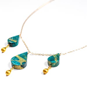May include: A gold chain necklace with three teardrop-shaped pendants. The pendants are made of a teal-colored material with gold accents.
