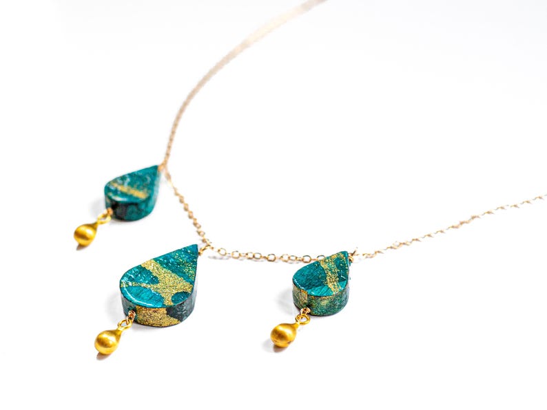 May include: A gold chain necklace with three teardrop-shaped pendants. The pendants are made of a teal-colored material with gold accents.