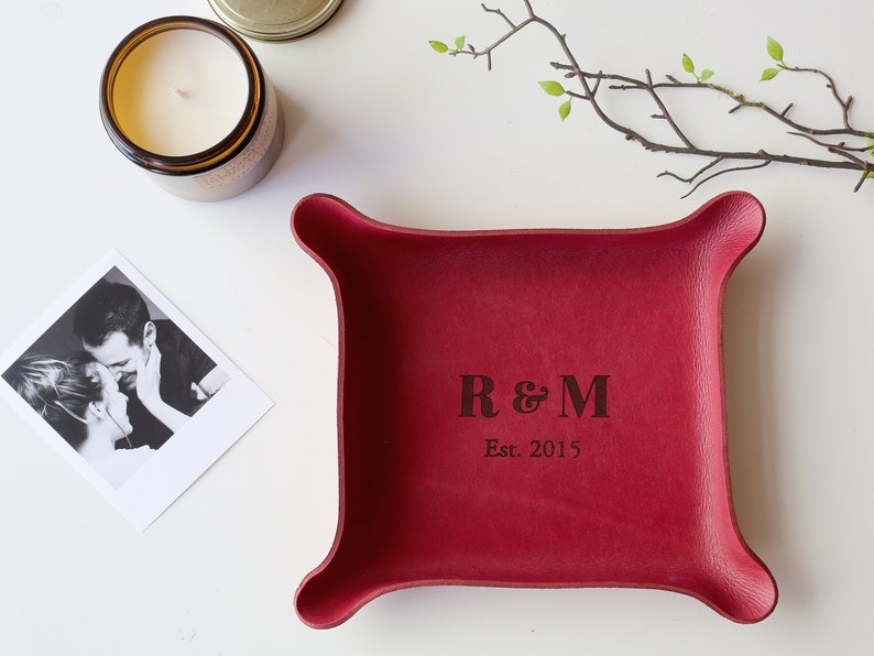May include: A burgundy leather tray with rounded corners, engraved with the initials "R & M" and the year "Est. 2015". A candle in a glass jar and a black and white photo are next to the tray. A sprig of greenery is in the background.