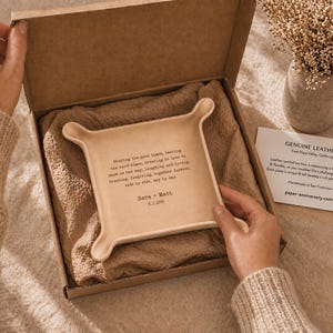 May include: A beige leather tray with rounded corners and a quote, presented in a cardboard box with a beige lining. The tray has a quote and the names "Sara + Matt" and a date. A card reads "Genuine Leather".