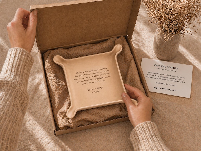May include: A beige leather tray with rounded corners and a quote, presented in a cardboard box with a beige lining. The tray has a quote and the names "Sara + Matt" and a date. A card reads "Genuine Leather".