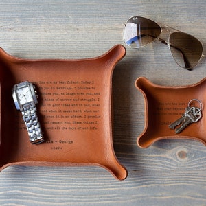May include: Three brown leather trays with rounded corners. The largest tray holds a silver watch. The medium tray has text and a set of keys. The smallest tray has text. Aviator sunglasses are on the right. Text on the trays.