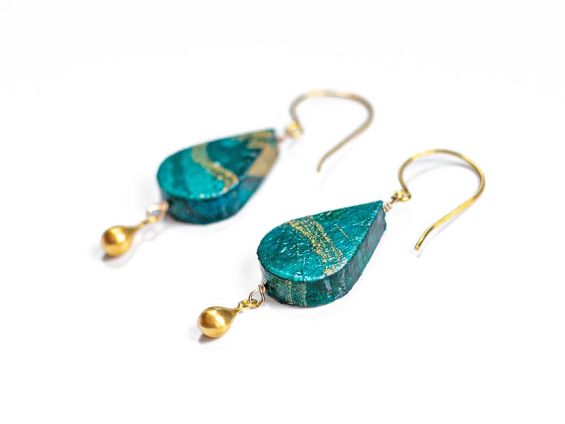 May include: Teal and gold teardrop shaped earrings with gold ball accents. The earrings are made of paper and have a unique, textured finish.