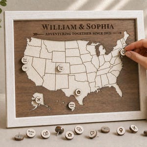 Puede incluir: Mapa de madera de los Estados Unidos con marco blanco, grabado con los nombres "William & Sophia" y el texto "Adventuring Together Since 2021." Incluye chinchetas para marcar ubicaciones. Una mano coloca un alfiler en forma de corazón.