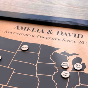 Peut inclure: Une carte en bois des &Eacute;tats-Unis avec des &eacute;tats en noir et blanc. La carte est encadr&eacute;e en noir et a une bordure de couleur cuivre. Le texte "Amelia & David Adventuring Together Since 2017" est grav&eacute; sur la bordure. Il y a plusieurs marqueurs en bois ronds avec du texte dessus, notamment "Next Trip", "Vacation", "We're Parents", "First Date", et "First Met".