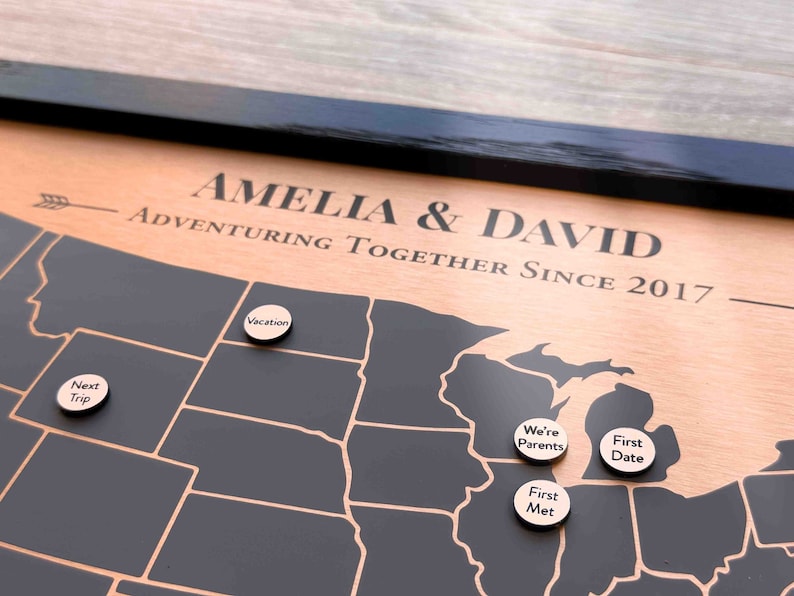 Peut inclure: Une carte en bois des &Eacute;tats-Unis avec des &eacute;tats en noir et blanc. La carte est encadr&eacute;e en noir et a une bordure de couleur cuivre. Le texte "Amelia & David Adventuring Together Since 2017" est grav&eacute; sur la bordure. Il y a plusieurs marqueurs en bois ronds avec du texte dessus, notamment "Next Trip", "Vacation", "We're Parents", "First Date", et "First Met".