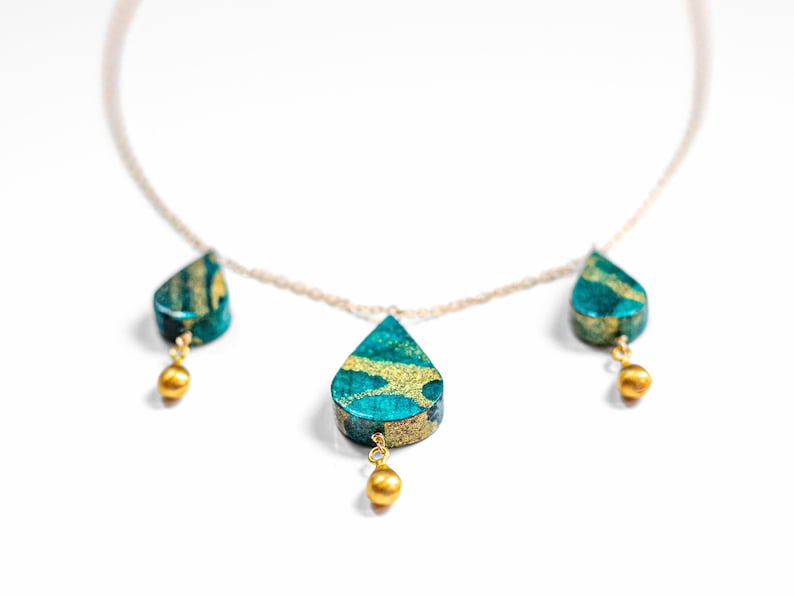 May include: A gold chain necklace with three teardrop-shaped pendants. The pendants are made of a green and gold material with a marbled pattern. Each pendant is accented with a small gold bead.