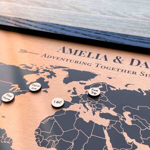 Peut inclure: Une carte du monde en bois avec un cadre noir et une surface de couleur cuivre. La carte est d&eacute;cor&eacute;e de petites &eacute;pingles en bois avec du texte comme "First Home", "Fell in Love", "I do!" et "First Trip". Le texte "Amelia & David Adventuring Together Since 20" est grav&eacute; sur la carte.