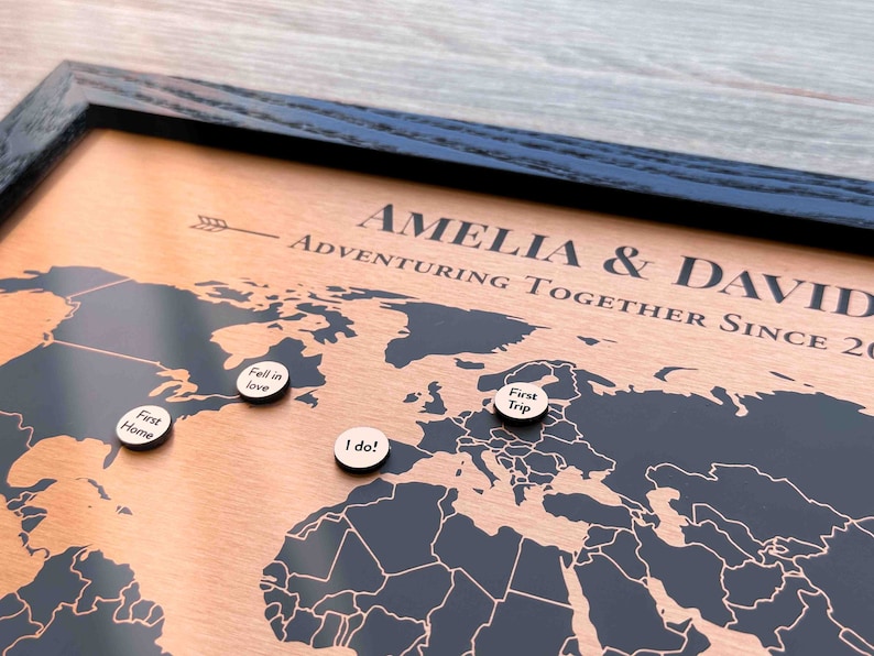 Peut inclure: Une carte du monde en bois avec un cadre noir et une surface de couleur cuivre. La carte est d&eacute;cor&eacute;e de petites &eacute;pingles en bois avec du texte comme "First Home", "Fell in Love", "I do!" et "First Trip". Le texte "Amelia & David Adventuring Together Since 20" est grav&eacute; sur la carte.