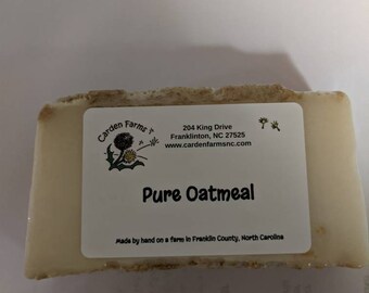 Può includere: Una saponetta bianca con un'etichetta marrone che recita "Garden Farms*" e "Pure Oatmeal". L'etichetta include anche l'indirizzo "204 King Drive, Franklinton, NC 27525" e il sito web "www.cardenfarmsnc.com". Il sapone è fatto a mano in una fattoria nella contea di Franklin, Carolina del Nord.