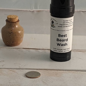 Puede incluir: Un pequeño tarro de cerámica marrón moteado con un tapón de corcho está junto a un tubo negro etiquetado "Best Beard Wash". El tubo contiene 71 gramos de producto. Una moneda está en primer plano.