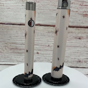 May include: Two white vape pens with black bat and spider designs. The pens are on black bases.
