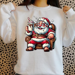 May include: White sweatshirt with a cartoon illustration of Santa Claus sitting in a chair, holding a mug of hot chocolate with marshmallows. The text "Merry & Bright" is printed on the sweatshirt.
