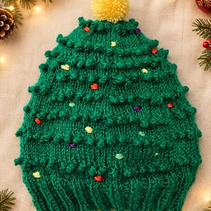 May include: A green knit beanie shaped like a Christmas tree, featuring a ribbed brim, textured bobble stitches throughout, and small colorful embroidered accents. The hat is topped with a single, round, yellow yarn pom-pom.