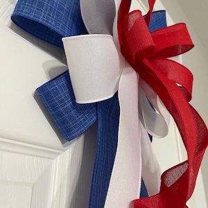 May include: A patriotic bow featuring blue, white, and red ribbons. The bow has a layered design with a textured blue ribbon, a smooth white ribbon, and a flowing red ribbon. The bow is likely used for decoration.
