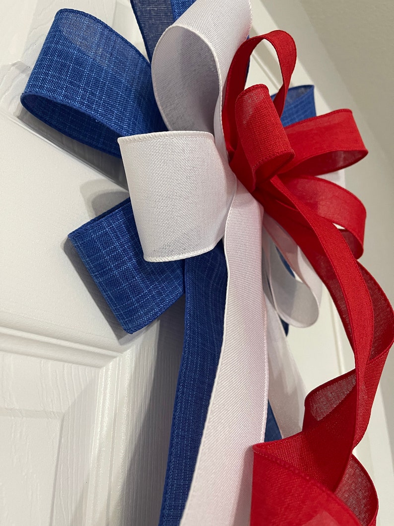 May include: A patriotic bow featuring blue, white, and red ribbons. The bow has a layered design with a textured blue ribbon, a smooth white ribbon, and a flowing red ribbon. The bow is likely used for decoration.