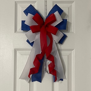 May include: A decorative bow in red, white, and blue, hanging on a white door. The bow features layers of blue patterned fabric, white sheer fabric, and a red ribbon. The bow is likely used for patriotic or holiday decor.
