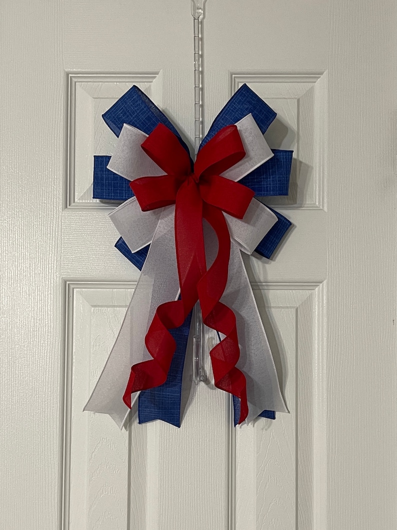 May include: A decorative bow in red, white, and blue, hanging on a white door. The bow features layers of blue patterned fabric, white sheer fabric, and a red ribbon. The bow is likely used for patriotic or holiday decor.